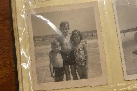 A phone photo of an old print, in an album: three children in swimsuits at the beach